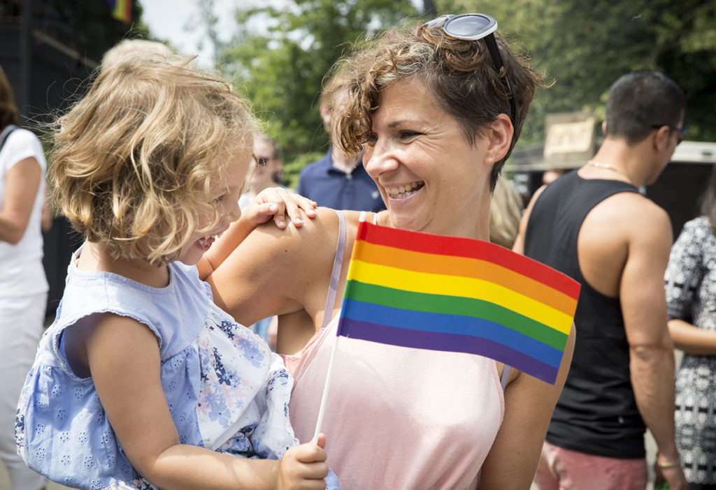 Mujer sonriente sostiene a una niña pequeña con una bandera arcoíris en un evento, simbolizando la diversidad y la inclusión.